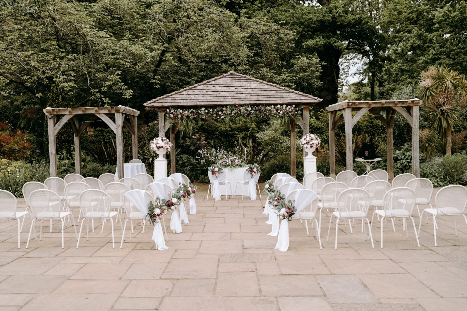 Outdoor wedding setup featuring rows of white chairs, floral arrangements, and a decorated arch in a lush garden.