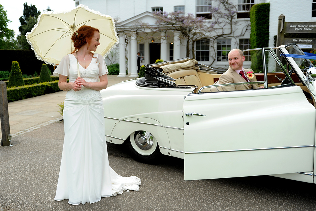 A woman in a white dress holding a parasol and looking over her shoulder at a man in a vintage car.
