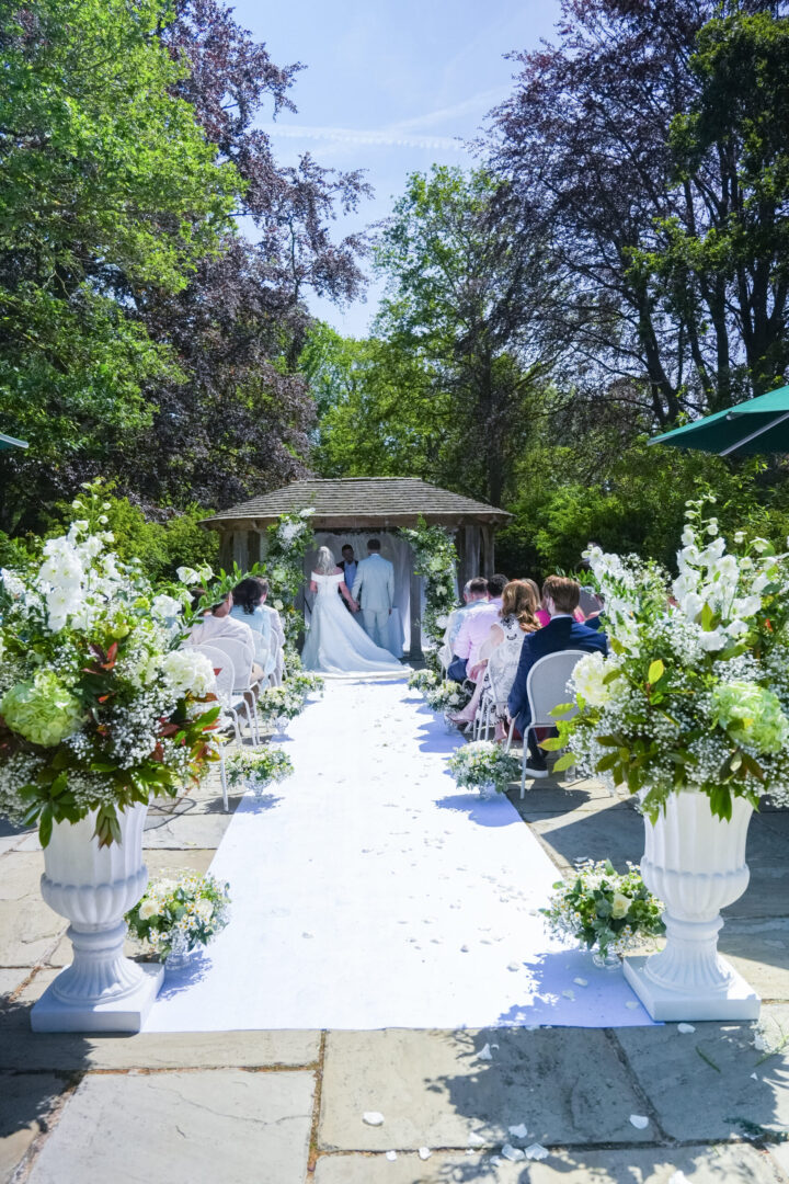 Outdoor wedding ceremony in a garden, featuring an arch adorned with flowers and guests seated on white chairs.