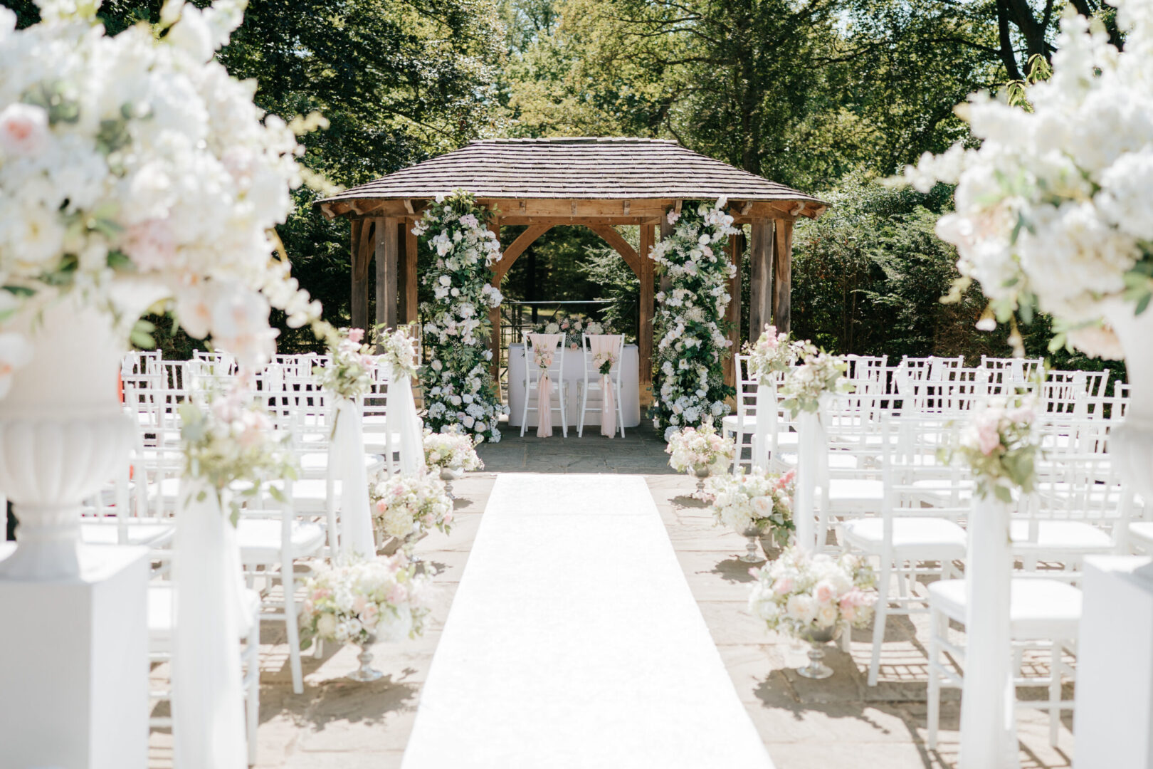 A large wooden wedding arch with rows of white chairs and a long white carpet on a sunny day.